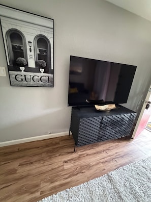 A flat-screen TV mounted on a textured black cabinet, a framed Gucci sign on the wall, and a wooden floor with a white rug.