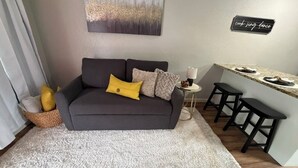Una sala de estar con un sofá gris, una alfombra blanca, una mesita auxiliar con una lámpara y una zona de cocina con un taburete de bar.