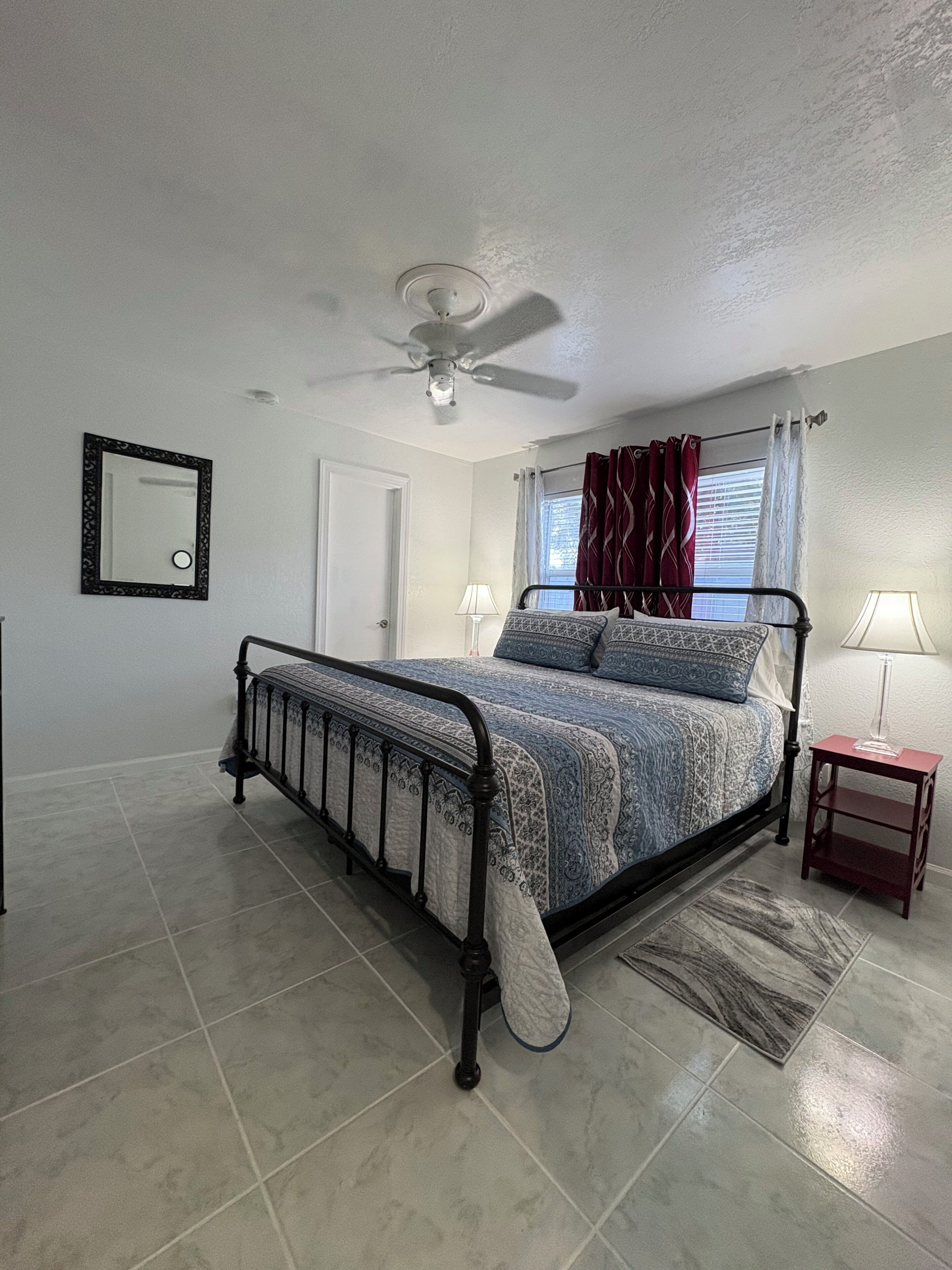 A bedroom with a bed, a ceiling fan, a window with curtains, a bedside table, and a mirror on the wall.