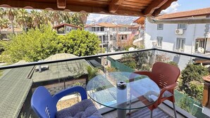 Un balcón con techo de madera, una mesa de vidrio, dos sillas y vistas a edificios vecinos y áreas verdes.
