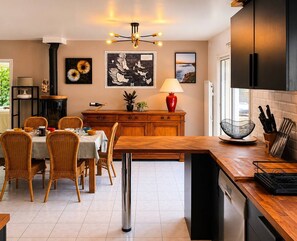 A modern kitchen with a dining area, featuring a wooden table, wicker chairs, a black stove, and a red vase. - Maison 8px, jacuzzi, jardin, 15 min à pied plage (QUIBERON)