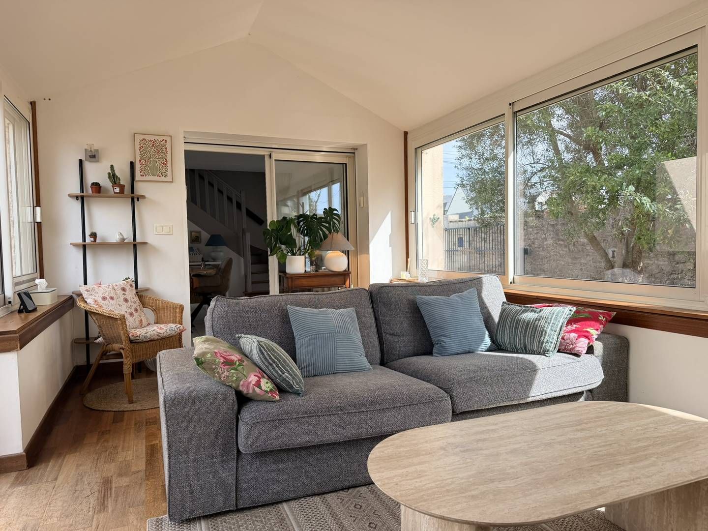 Un salon moderne avec un canapé gris, une table basse en bois et une vue sur l’extérieur grâce à de grandes fenêtres.