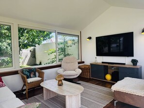 A living room with a sofa, armchair, coffee table, TV, and a large window with a view of greenery. - Maison 8px, jacuzzi, jardin, 15 min à pied plage (QUIBERON)