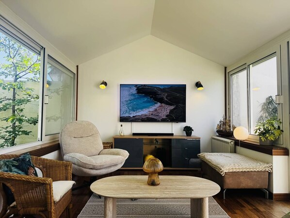 A modern living room with a large flat-screen TV, a comfortable armchair, a small table, and a bench. - Maison 8px, jacuzzi, jardin, 15 min à pied plage (QUIBERON)