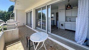 Un balcón con mesa y sillas, un ventanal corredero que da a una cocina y vistas a árboles.