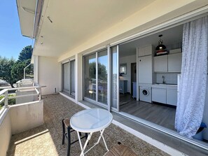Ein Balkon mit Tisch und Stühlen, eine Glastür, die in eine Küche führt, und Blick auf Bäume.