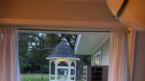 Blick durch ein Fenster auf einen kleinen Pavillon im Garten, einen grünen Container und eine Klimaanlage.