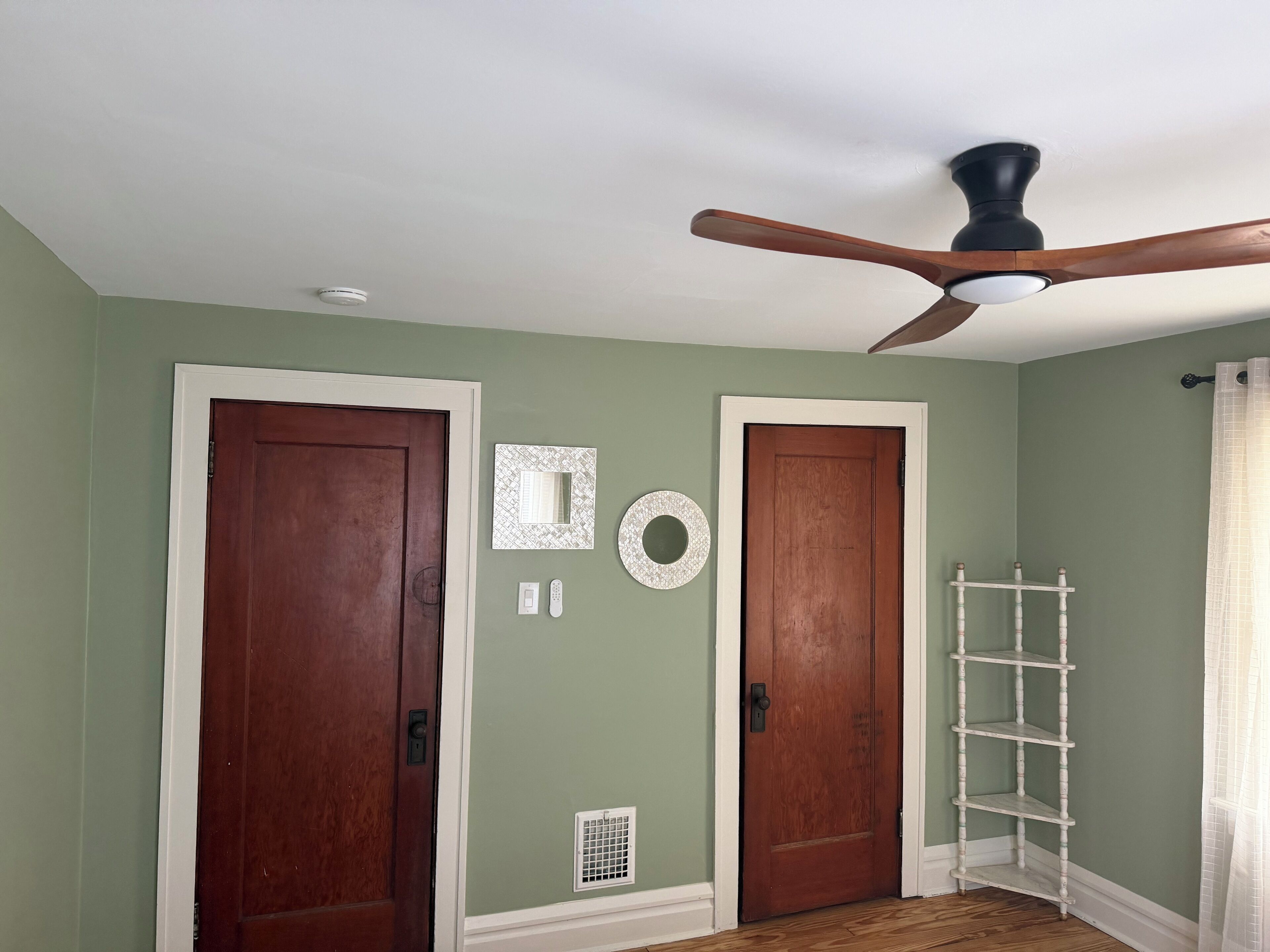 A room with two wooden doors, a ceiling fan, a white shelf, and a vent on the wall.