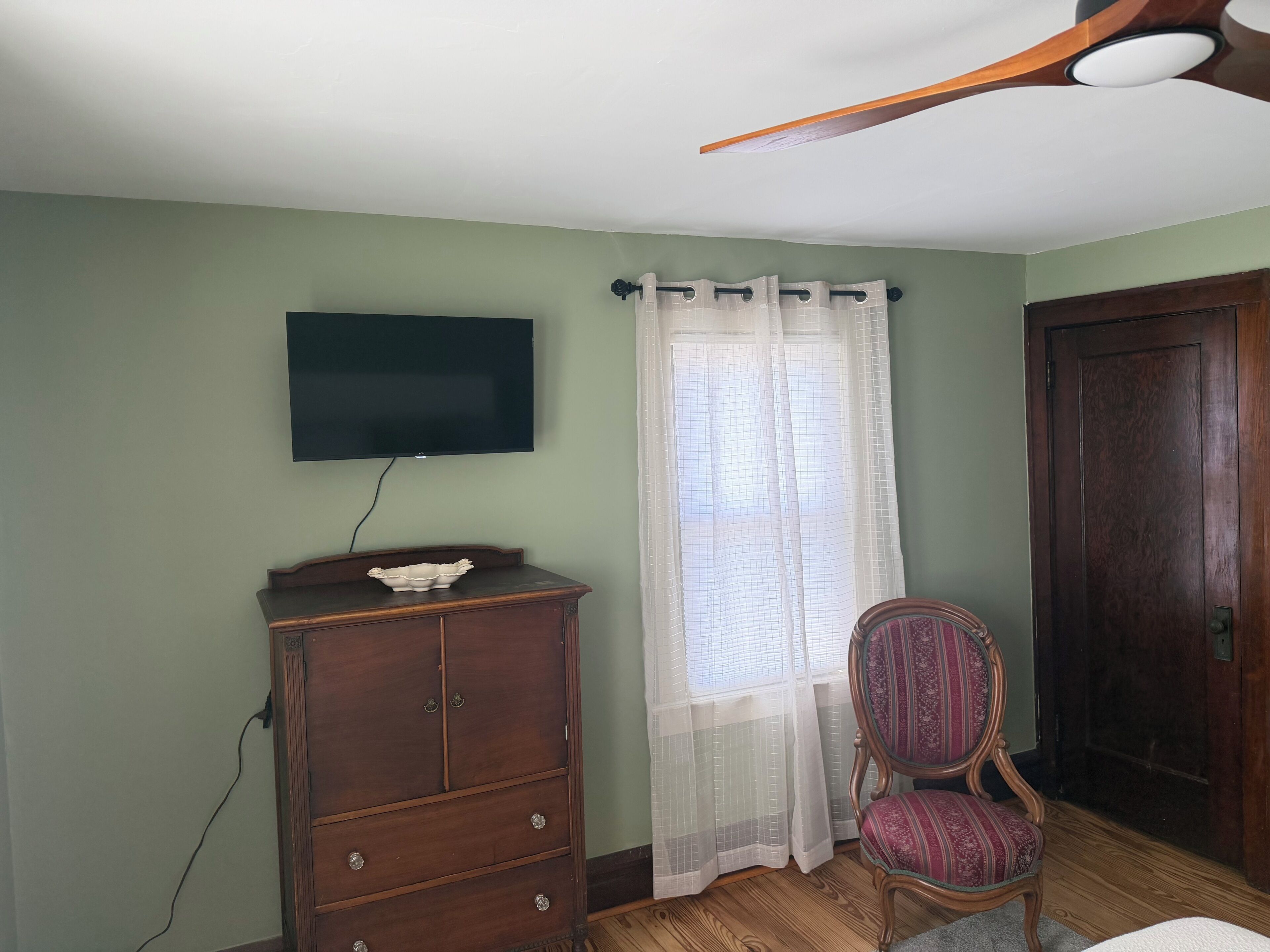 Una habitació amb un armari de fusta, televisió penjada a la paret, una cadira, una finestra amb cortines transparents i una porta.