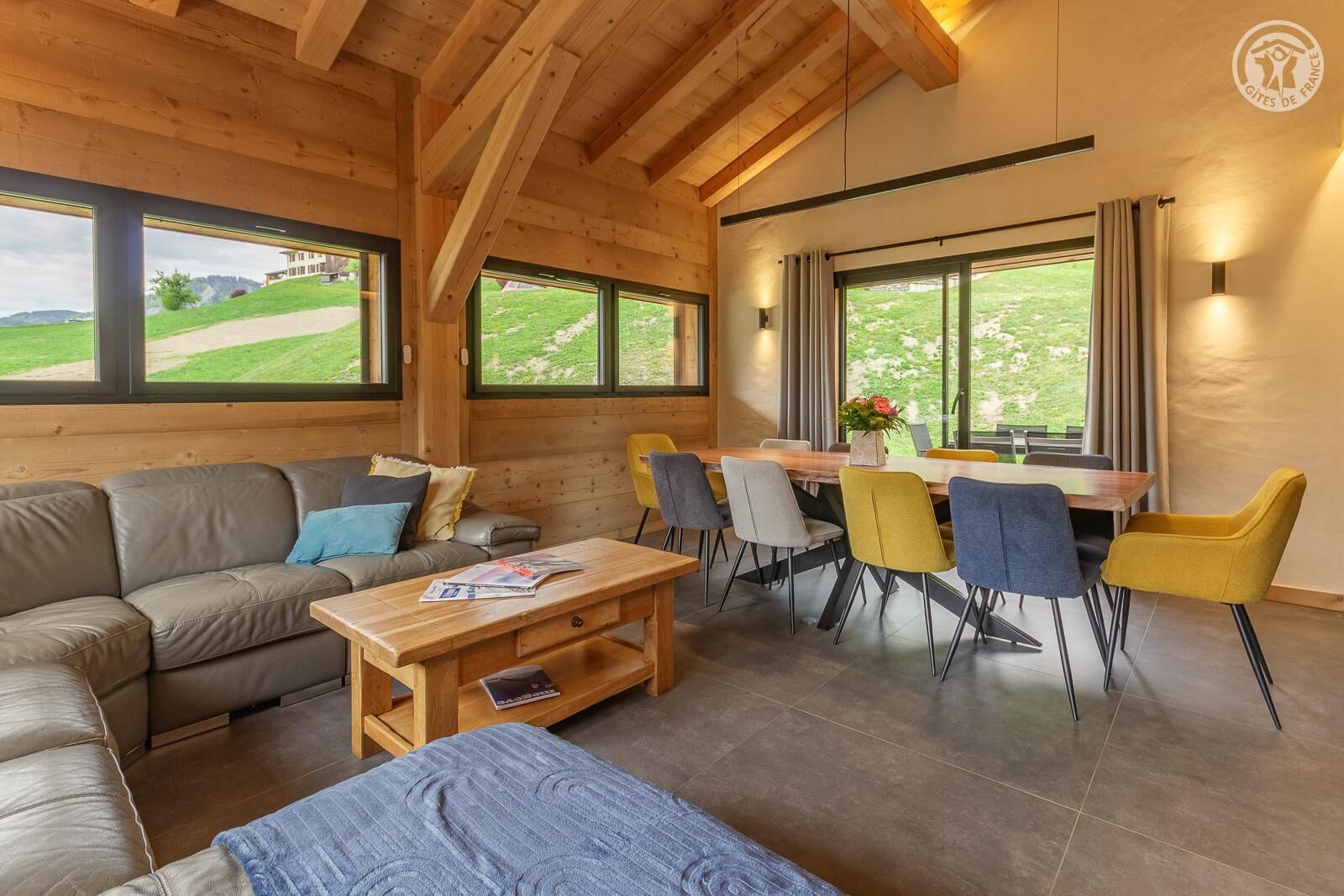 A wooden interior with a sofa, dining table, chairs, and large windows.