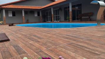 A modern house with a swimming pool, a patio with wooden flooring, and a garden with purple flowers.