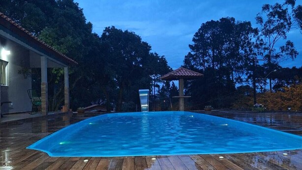 A swimming pool with a wooden deck, surrounded by trees and a house. - Sitio toa toa 2 (São roque)