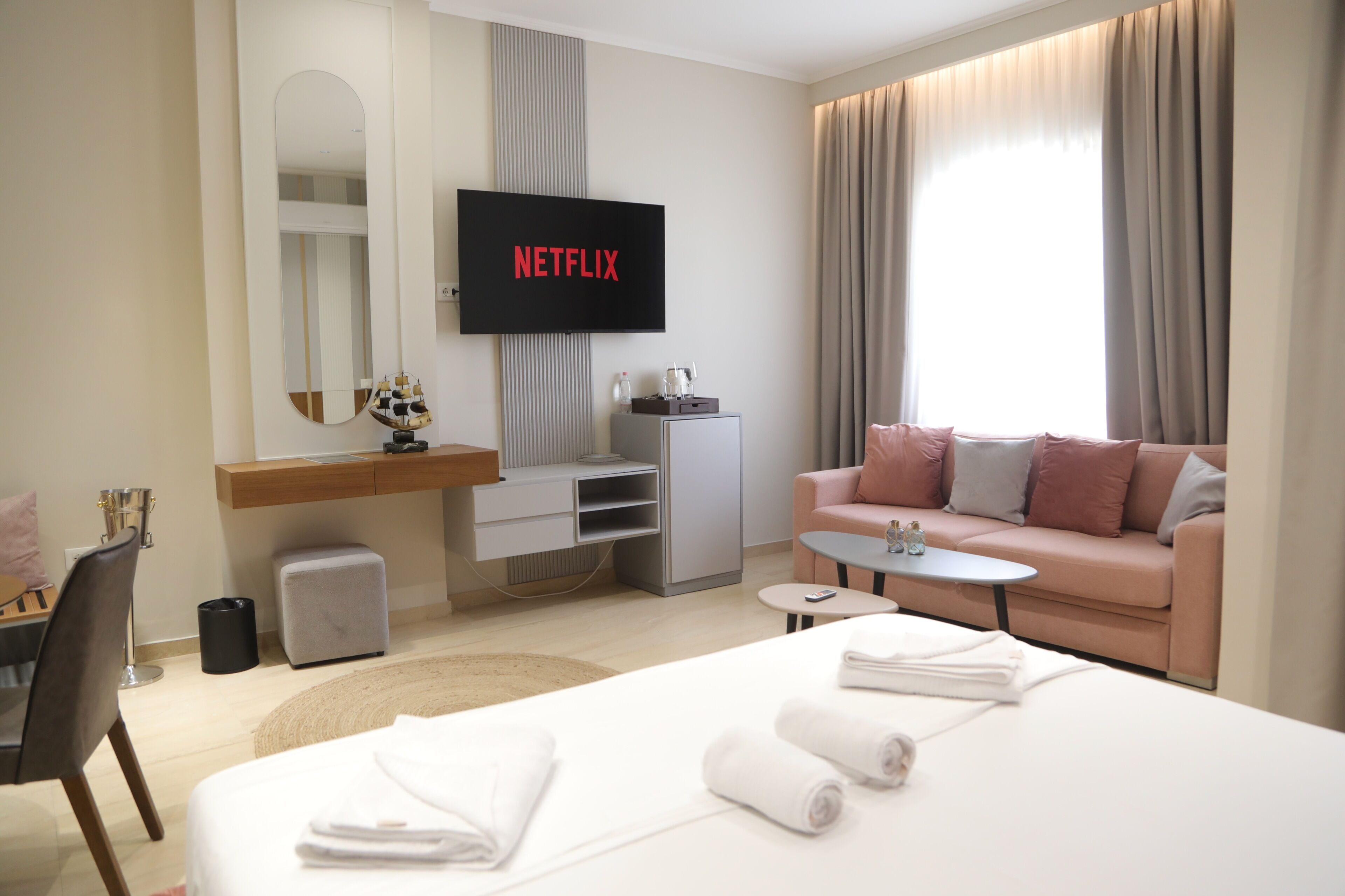 A modern hotel room with a bed, a sofa, a TV displaying 'NETFLIX,' a desk, and a small table.