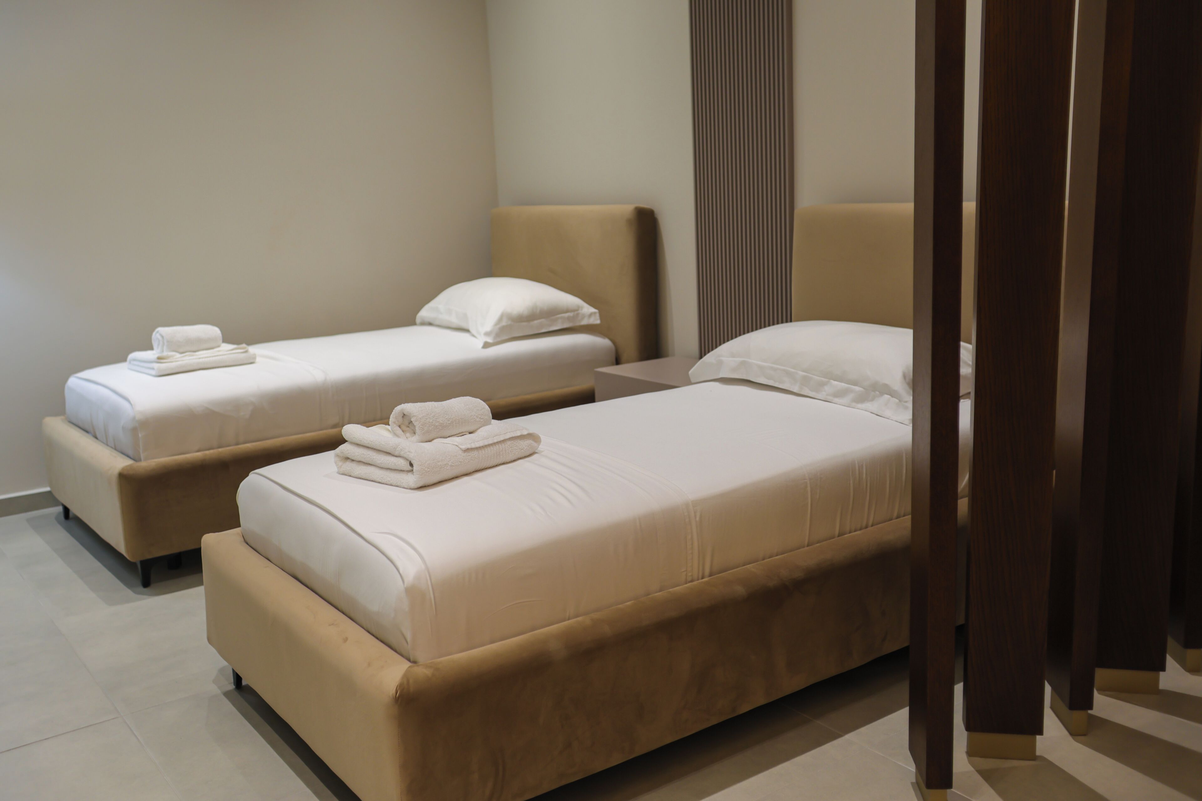 Two beds with white linens and beige headboards in a room with a wooden floor.