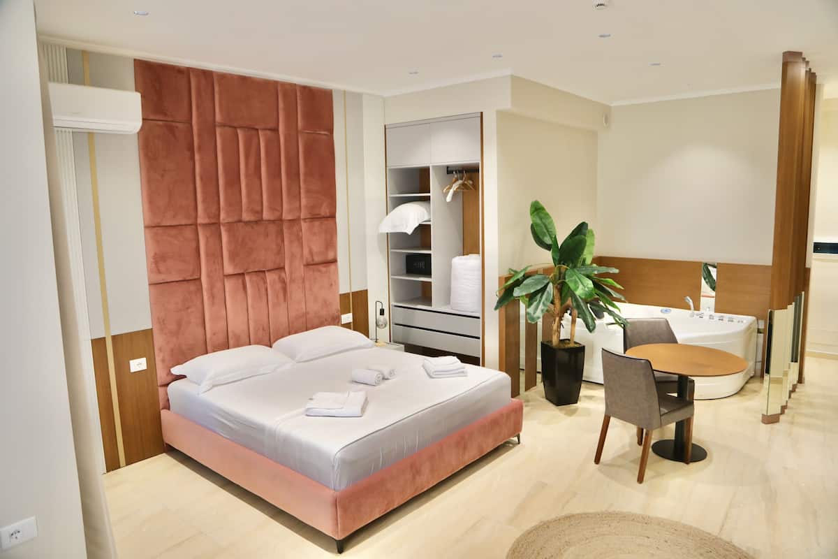 A modern hotel room with a large bed, a seating area, a built-in wardrobe, a plant, and a bathtub.