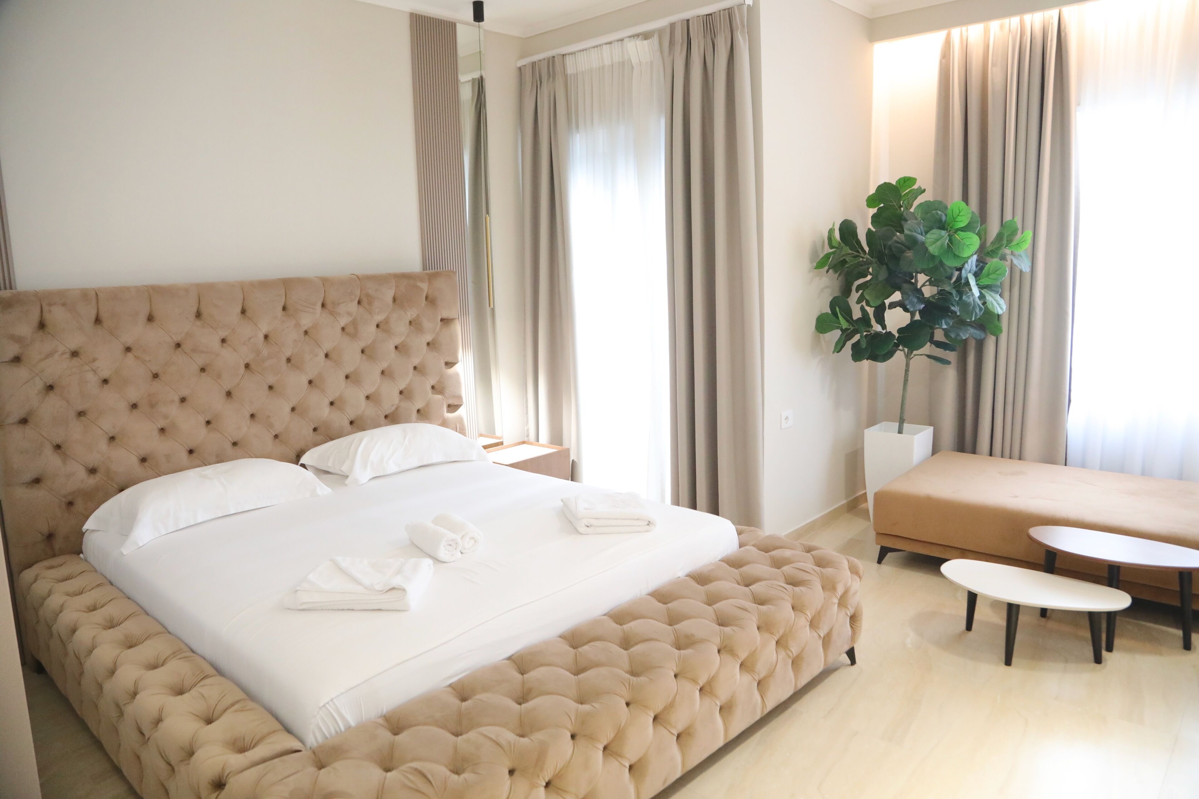 Ein großes Bett mit einem gebügelten Kopfteil, ein Nachttisch, eine Couch und ein Topf mit Pflanze in einem modernen Hotelzimmer.