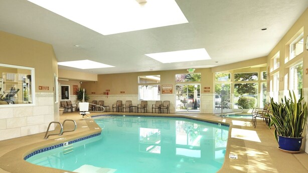An indoor swimming pool with clear water, surrounded by beige walls and a tiled floor. There are chairs and tables around the pool, and a potted plant is visible to the right. - 3 Modern Units | Explore Downtown Attractions | Free Breakfast, Parking, & Pool (Salt Lake County)