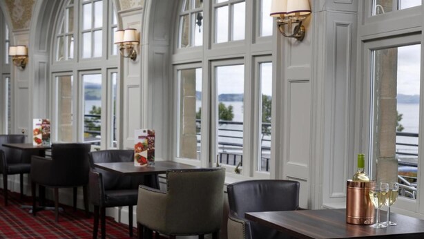 A restaurant with a red plaid carpet, tables with chairs, and a view of a body of water through large windows. - Best Western Argyll Hotel (Dunoon)