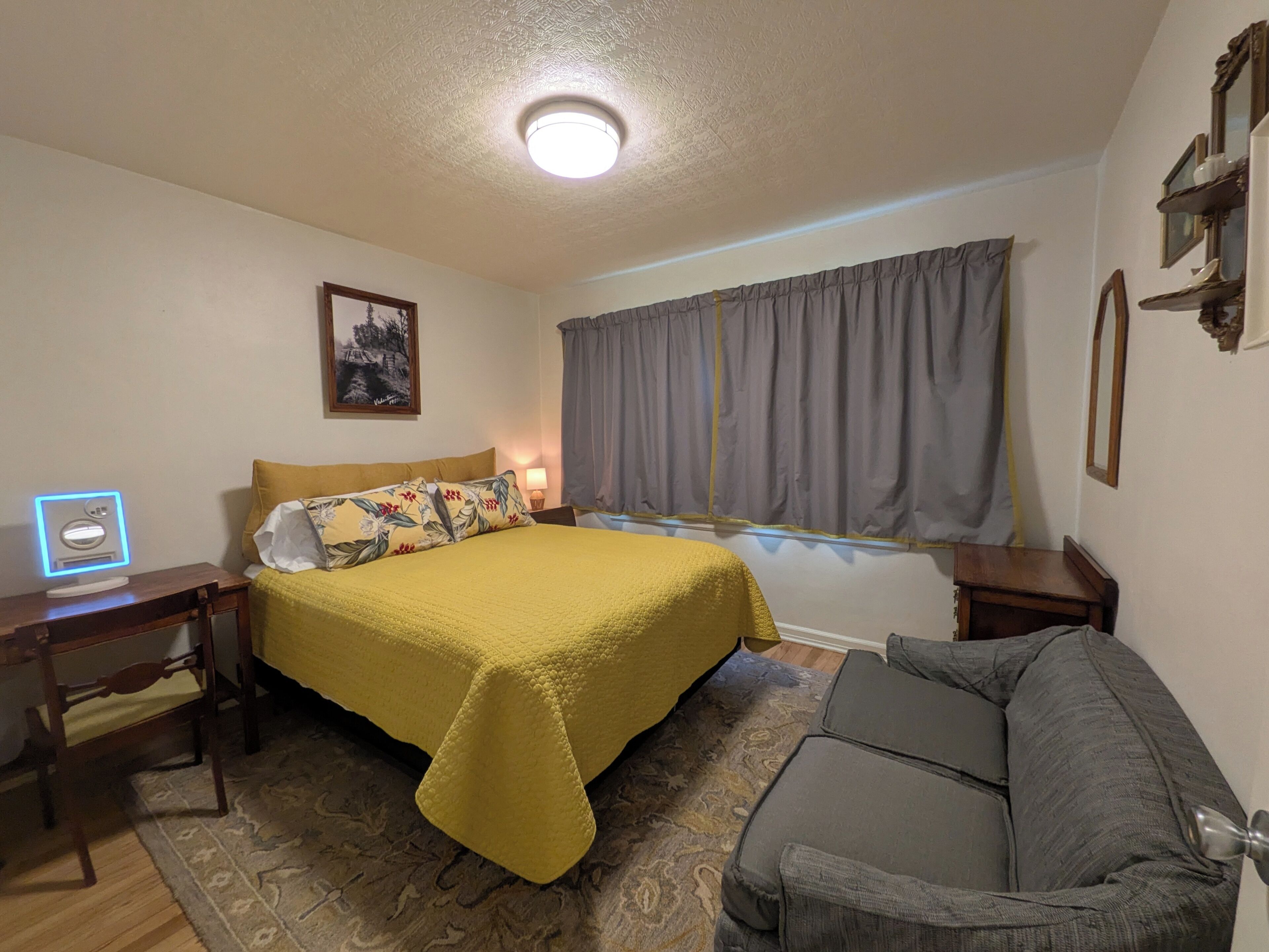 Ein Schlafzimmer mit Bett, Nachttisch, Lampe, einem Fenster mit Vorhängen, einer Couch und einem Wandregal.