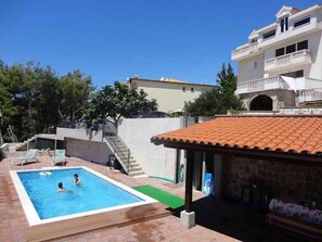 Ein Schwimmbecken mit zwei Personen, einer überdachten Terrasse und einem zweistöckigen Gebäude mit Balkonen.