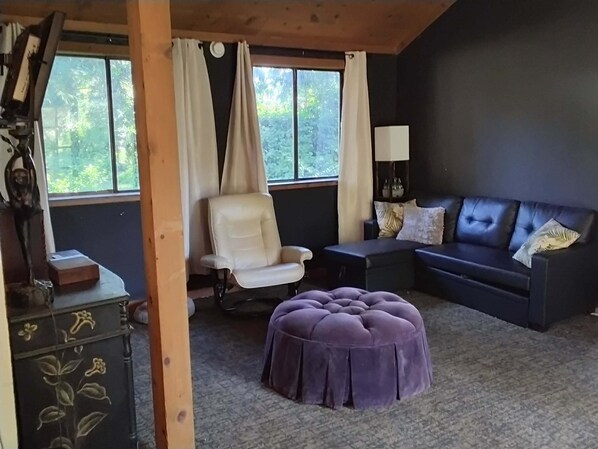 A room with a dark-colored sofa, a white chair, a purple ottoman, a lamp, and a window with curtains.