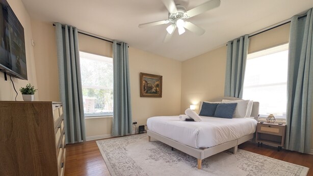 A bedroom with a bed, a ceiling fan, a nightstand, a desk, and a window with curtains. - Riverside 2BR | Movie Night Stay (Jacksonville)