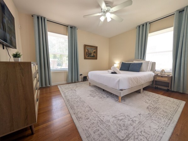 A bedroom with a bed, a ceiling fan, a nightstand, a desk, and a window with curtains. - Riverside Retreat 2BR w/ Big Screen Projector (Jacksonville)