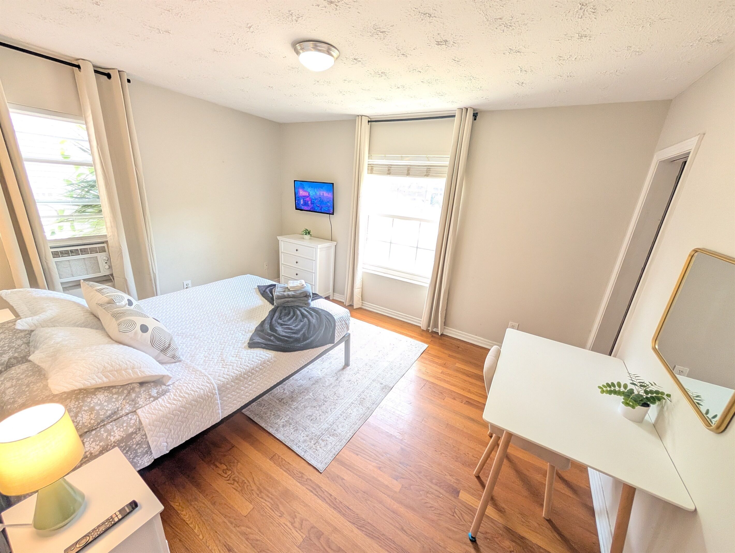 A bedroom with a bed, bedside table, lamp, TV, and a small desk with a mirror.