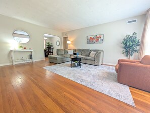 Ein Wohnzimmer mit Holzdielenboden, einem grauen Ecksofa, einem Couchtisch, einem runden Spiegel und einer Topfpflanze.
