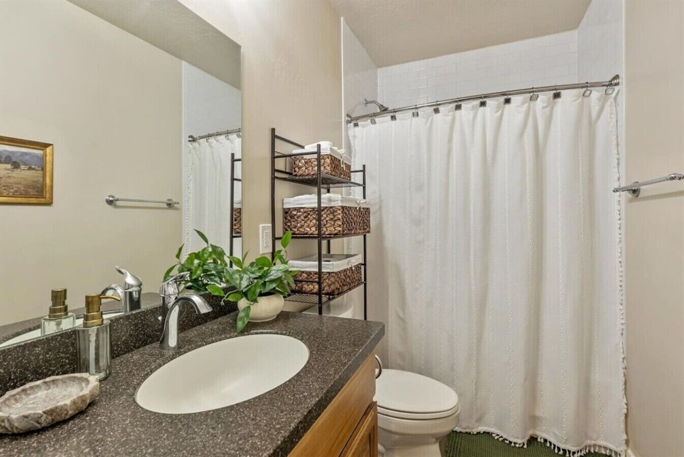 A bathroom with a granite countertop sink, a mirror, a toilet, a shower curtain, and a shelf with baskets.