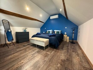 A bedroom with a sloped ceiling, a bed with blue bedding, a white bench, a dresser, and a mirror stand.