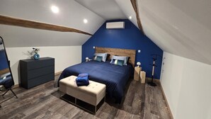 A bedroom with a sloped ceiling, a bed with multiple pillows, a bedside table, a dresser, a bench, and a floor lamp.