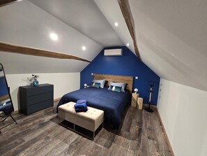 A bedroom with a sloped ceiling, a bed with multiple pillows, a bedside table, a dresser, a bench, and a floor lamp.