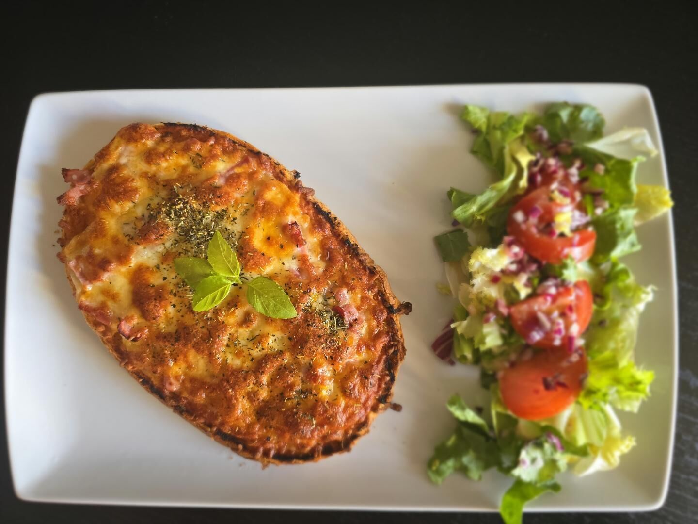 A rectangular plate with a food item topped with cheese and herbs, and a side salad with lettuce, tomatoes, and other greens.