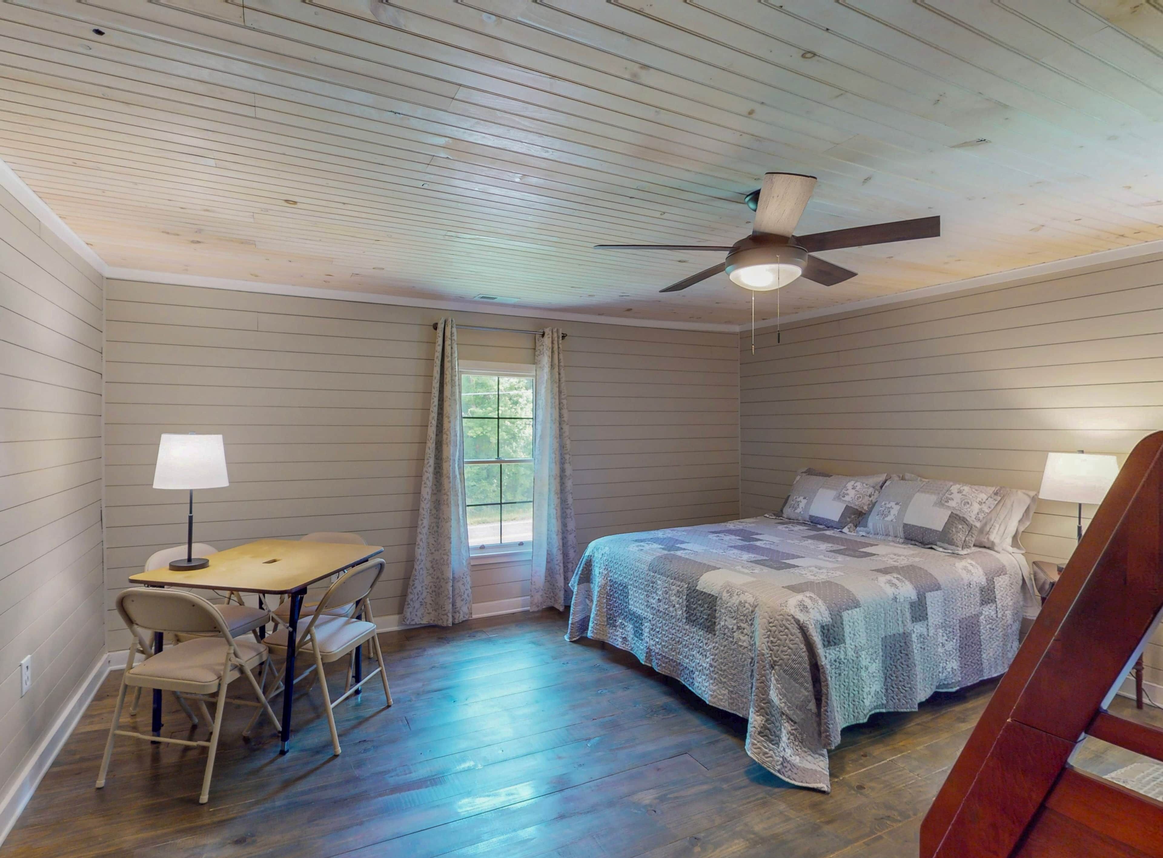 A bedroom with a bed, a desk with a chair, a ceiling fan, and a window with curtains.