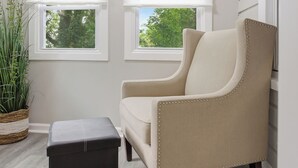 Ein Zimmer mit einem beigen Sessel, einem schwarzen Hocker und zwei Fenstern mit Jalousien.