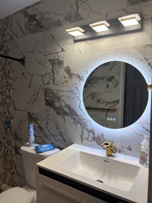 A bathroom with a marble wall, a round mirror with a light, a white sink, and a toilet.