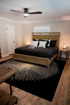 Ein Schlafzimmer mit einem Bett, einem Deckenventilator, Nachttischlampen, einem Holzkopfteil, einem schwarz-weißen Teppich und einer Holzbank.