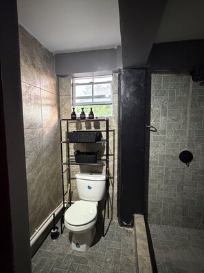 A compact bathroom with a toilet, a shelf with toiletries, a window, and a shower area. - Cozy 2-Bedroom Apartment Near Airport & Beaches in Vieux Fort (Vieux Fort)