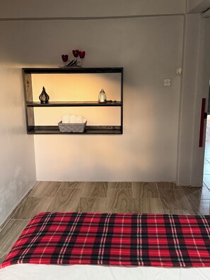 Une pièce avec un plancher en bois, une couverture à carreaux rouges sur le lit, une étagère murale ornée d’objets décoratifs et une porte avec une poignée rouge.