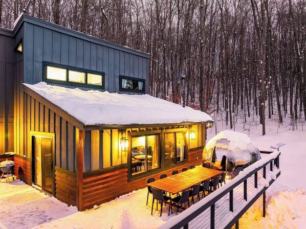 Una cabaña moderna con comedor y ventanas amplias, rodeada de nieve y árboles.