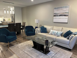 A modern living room with a glass coffee table, blue armchairs, a beige sofa with patterned cushions, a dining area with wooden chairs, and a kitchen with white cabinets and pendant lighting.