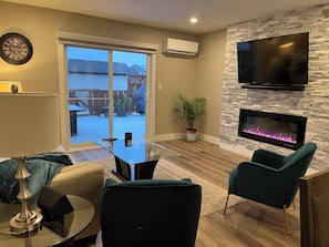 A modern living room with a glass coffee table, teal chairs, a fireplace, a TV, and a sliding glass door leading to a deck.