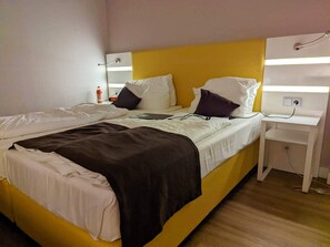 A double bed with white and brown bedding, a bedside table with a lamp, and a power outlet.