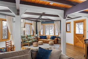 A cozy cabin interior with a wooden ceiling, a sofa, chairs, and a large window with a view of a snowy forest. - Retro Treehaus: 3 Kings, Bunks & Treetop Hot Tub (Bellaire)