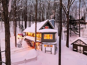 A two-story cabin with a snow-covered roof, a visible chimney, and a lit interior. - Retro Treehaus: 3 Kings, Bunks & Treetop Hot Tub (Bellaire)