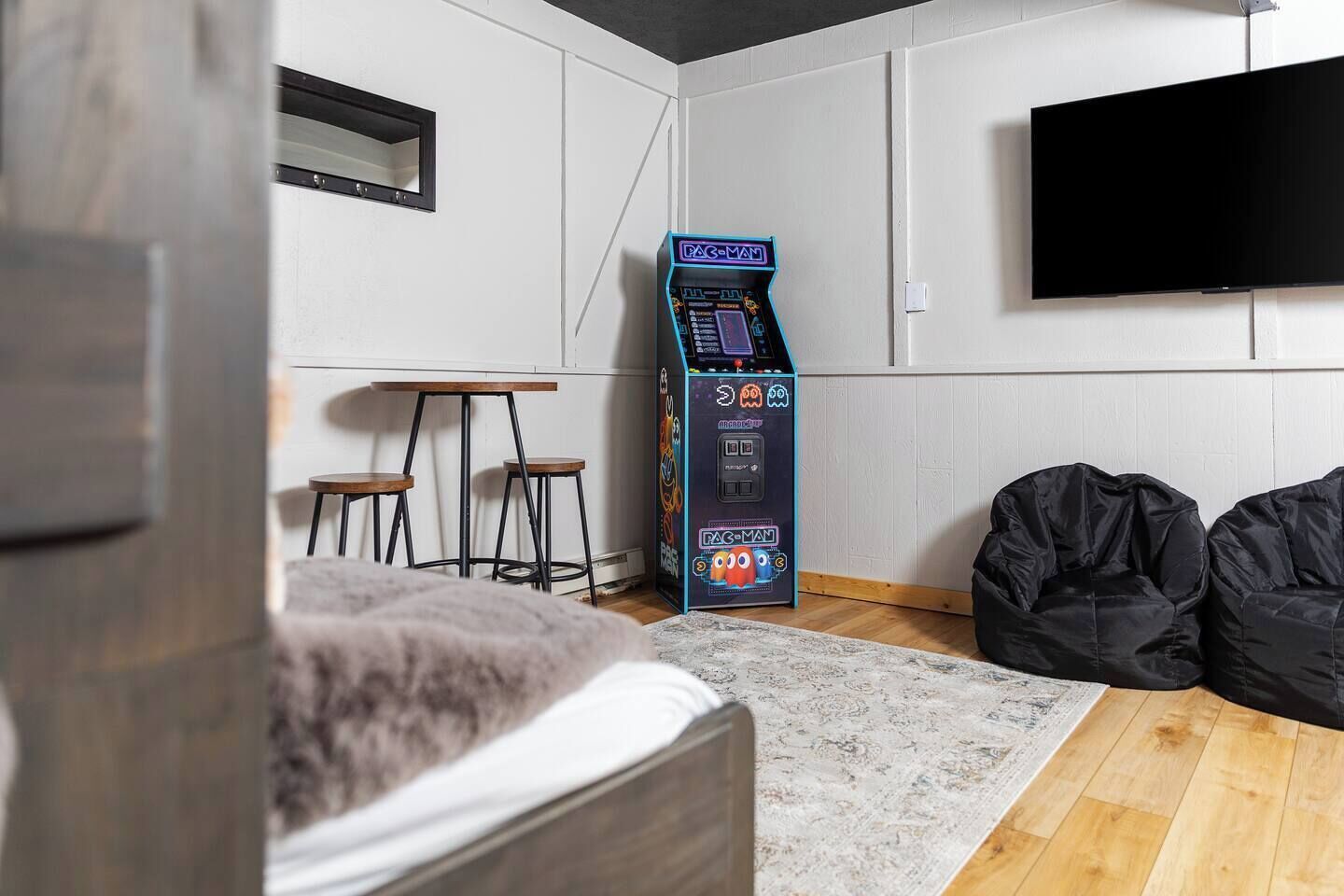 Ein Zimmer mit einem Bett, einer Pac-Man-Spielmaschine, zwei Barhocker, einem Flachbildfernseher und schwarzen Sitzsäcken.
