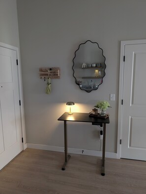 Ein Zimmer mit Schreibtisch, einem an der Wand befestigten Spiegel, einer kleinen Ablage mit Dekoration und einer Tür.