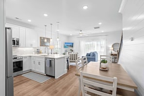 A modern kitchen with white cabinetry, a central island, and a dining area with a wooden table and chairs. - Walk to Beach | Brand New Luxury Home Myrtle Beach (Myrtle Beach)