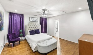 Ein Schlafzimmer mit einer karuppgebügelten Kopfteile, einem Bett mit weißer Bettwäsche, einem grauen Pouf, einem violetten Sessel und violetten Vorhängen.